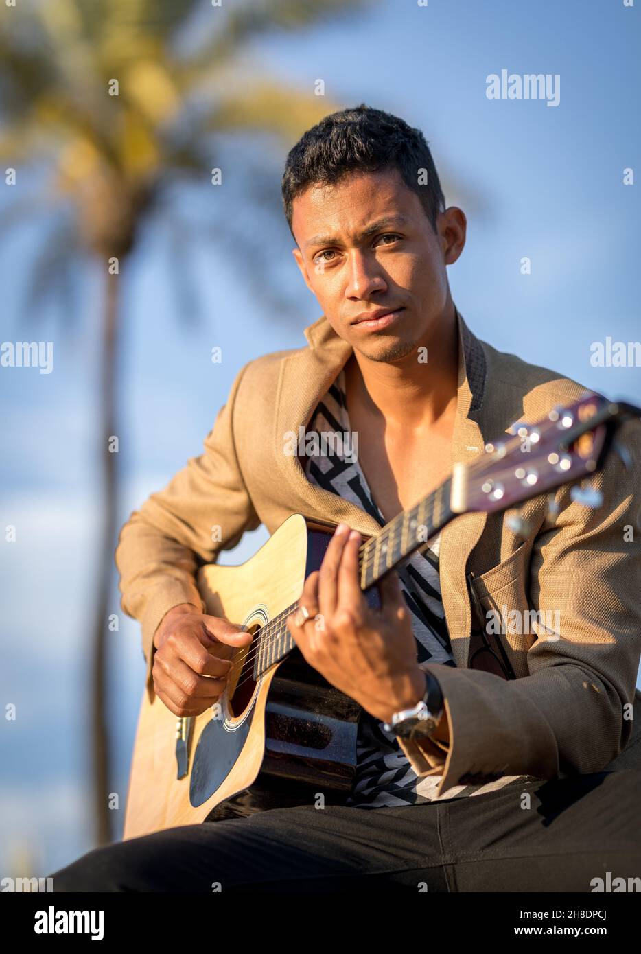 Portrait of Latin man looking at camera and playing guitar on the street with palm trees in the background at sunset Stock Photo