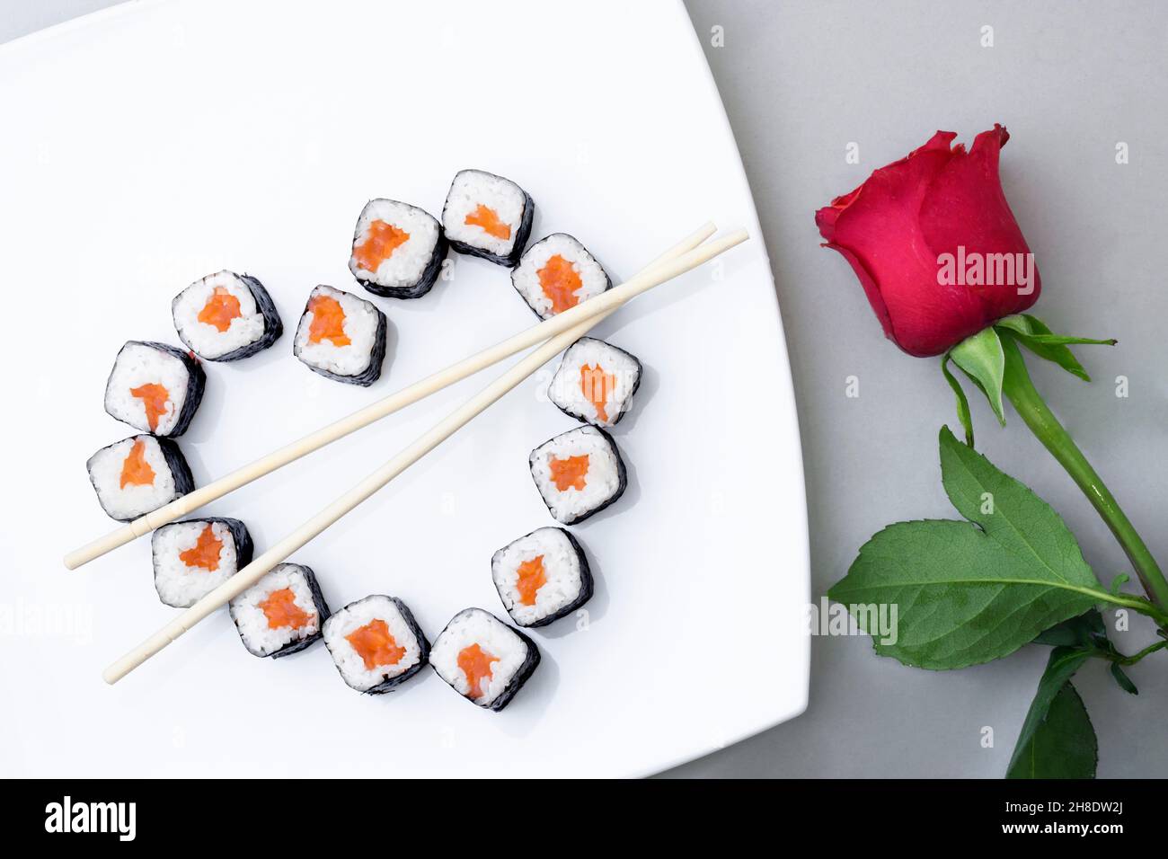 Sushi rolls laid out in the shape of a heart on a white plate. Red rose on a gray background. Creative greetings. The concept of Japanese cuisine for Stock Photo