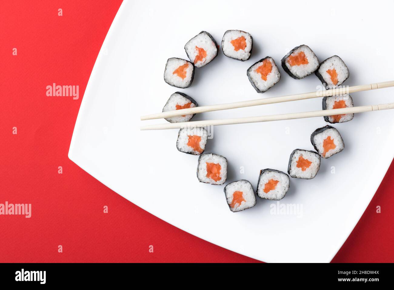 Sushi rolls laid out in the shape of a heart on a white plate, on a red background. Creative greetings. The concept of Japanese cuisine for Valentine' Stock Photo