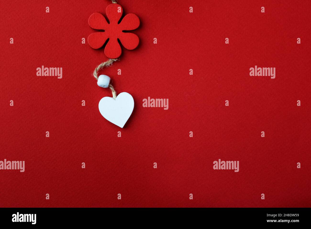 Composition background for Valentine's Day. A white heart made of wood hangs on a natural thread under a red flower, on a red background. Copy space Stock Photo