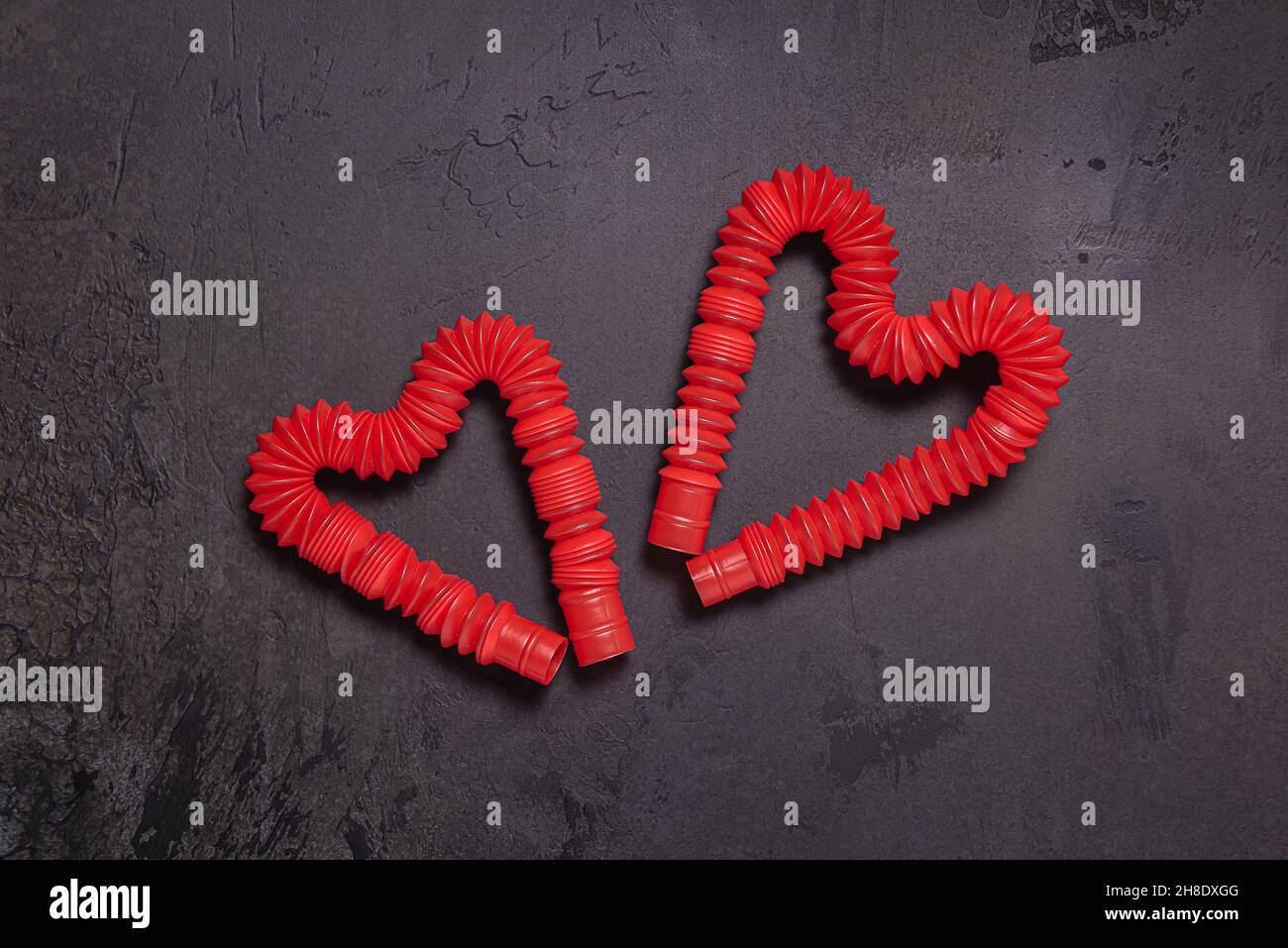 Two stretched red pop it tube children toys bent in shape of hearts. Plastic antistress bright toys on black textured stone background. Flat lay. Copy Stock Photo