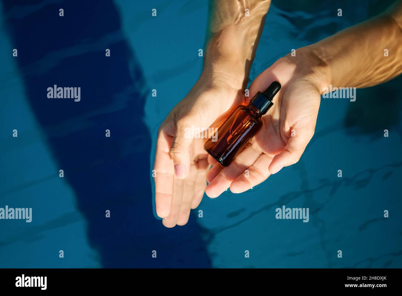 Woman hand holding empty organic serum skincare product glass amber bottle for use in a daytime with plain light blue swimming pool water background.  Stock Photo