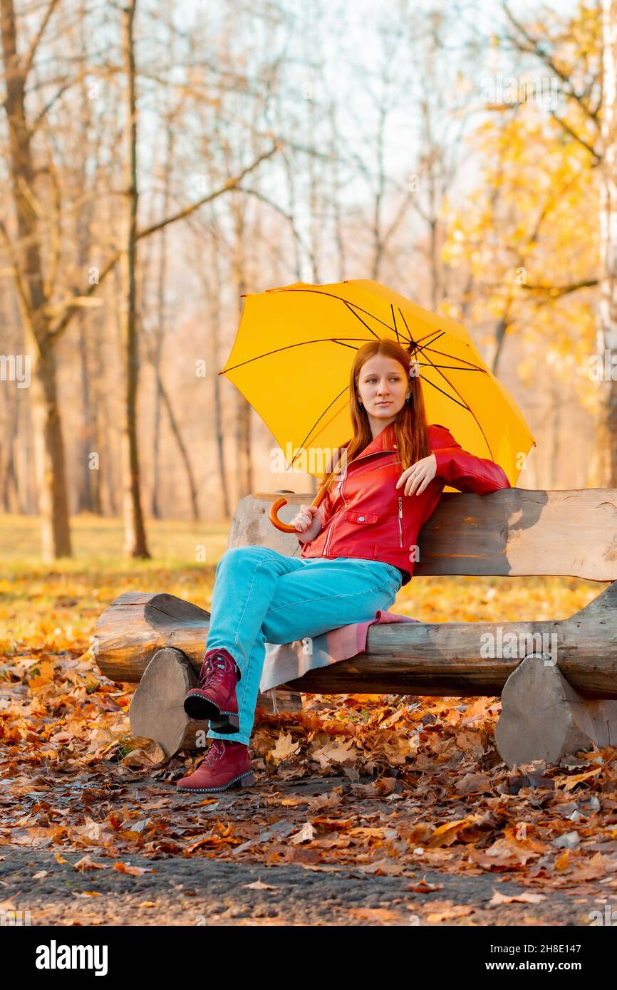 A girl alone sits under an umbrella in an autumn park on a bench cut from a log Stock Photo