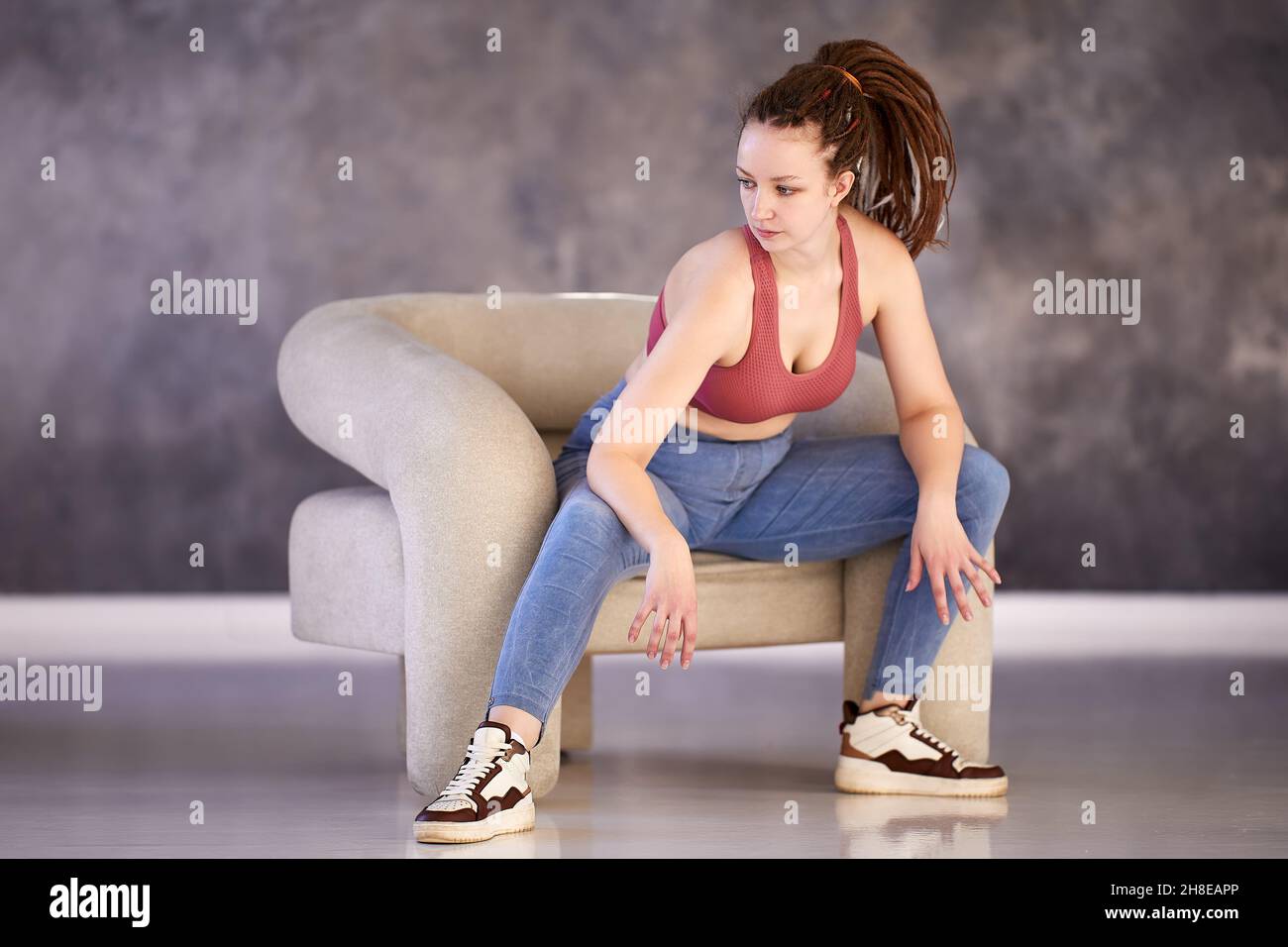 Woman with box braids dressed in tank top and jeans sits in chair thinking. Stock Photo