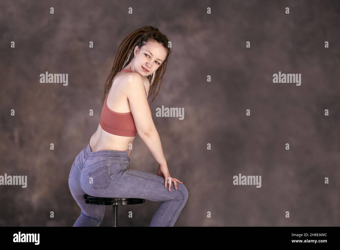 Spiral bar stool on which young white woman sits in tank top and jeans with her back half turned. Stock Photo