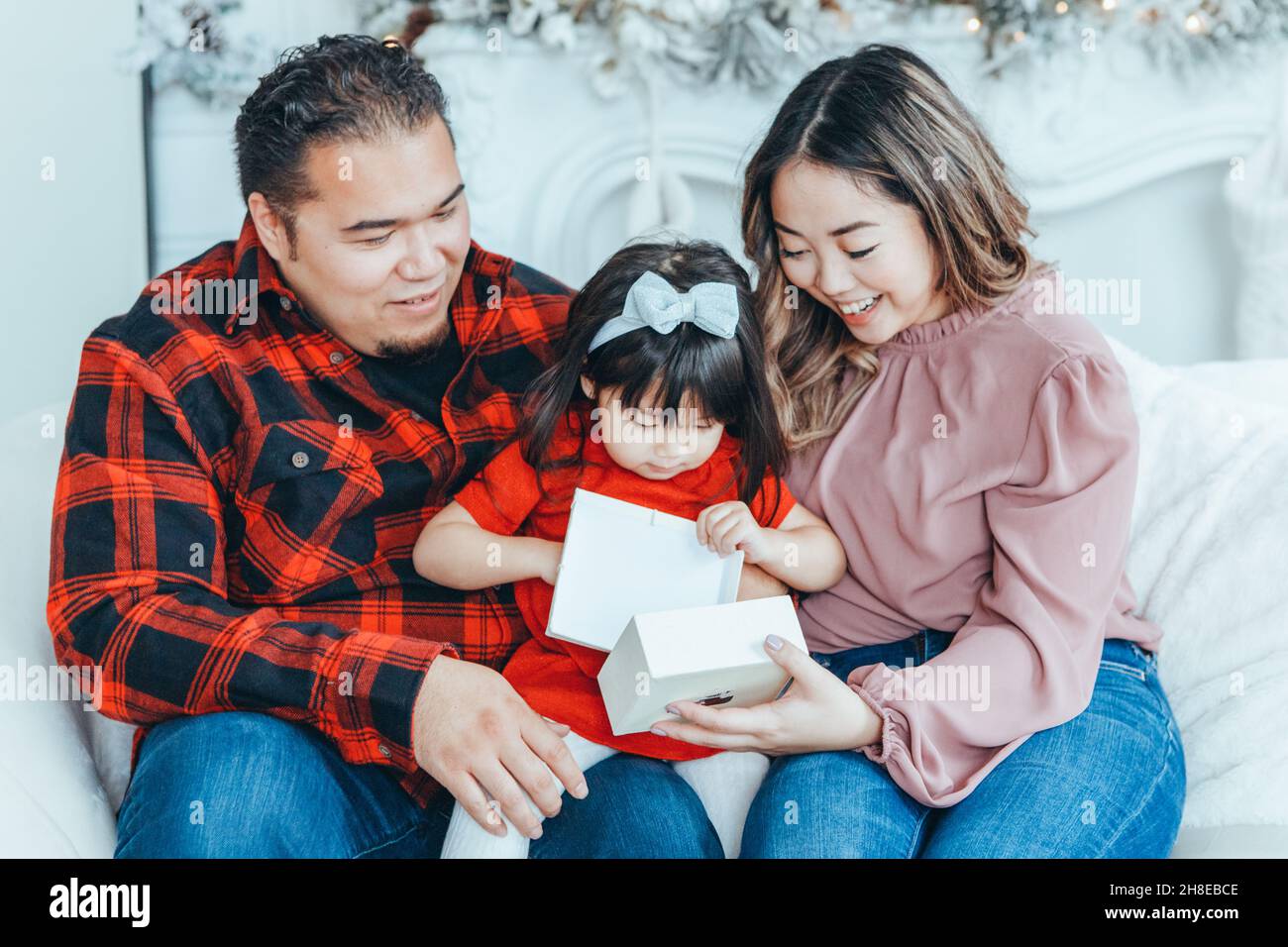 Asian family father, mother giving gift box with present to daughter toddler girl celebrating Christmas or New Year. Mixed race mom, dad, daughter sit Stock Photo
