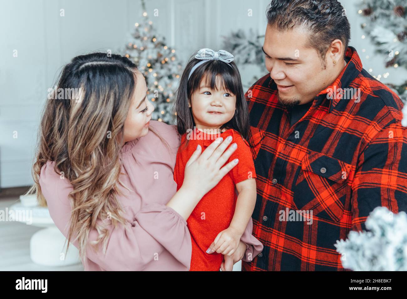 Asian family father, mother with daughter toddler girl celebrating Christmas or New Year. Mixed race mom, dad, daughter decorating Christas tree huggi Stock Photo