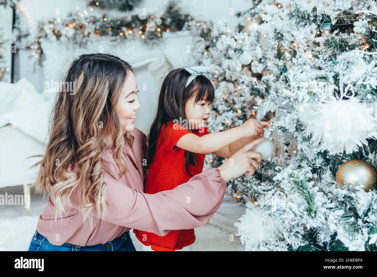 Asian family mother with daughter toddler girl celebrating Christmas or New Year. Mixed race mom, daughter decorating Christmas tree at home. Winter h Stock Photo