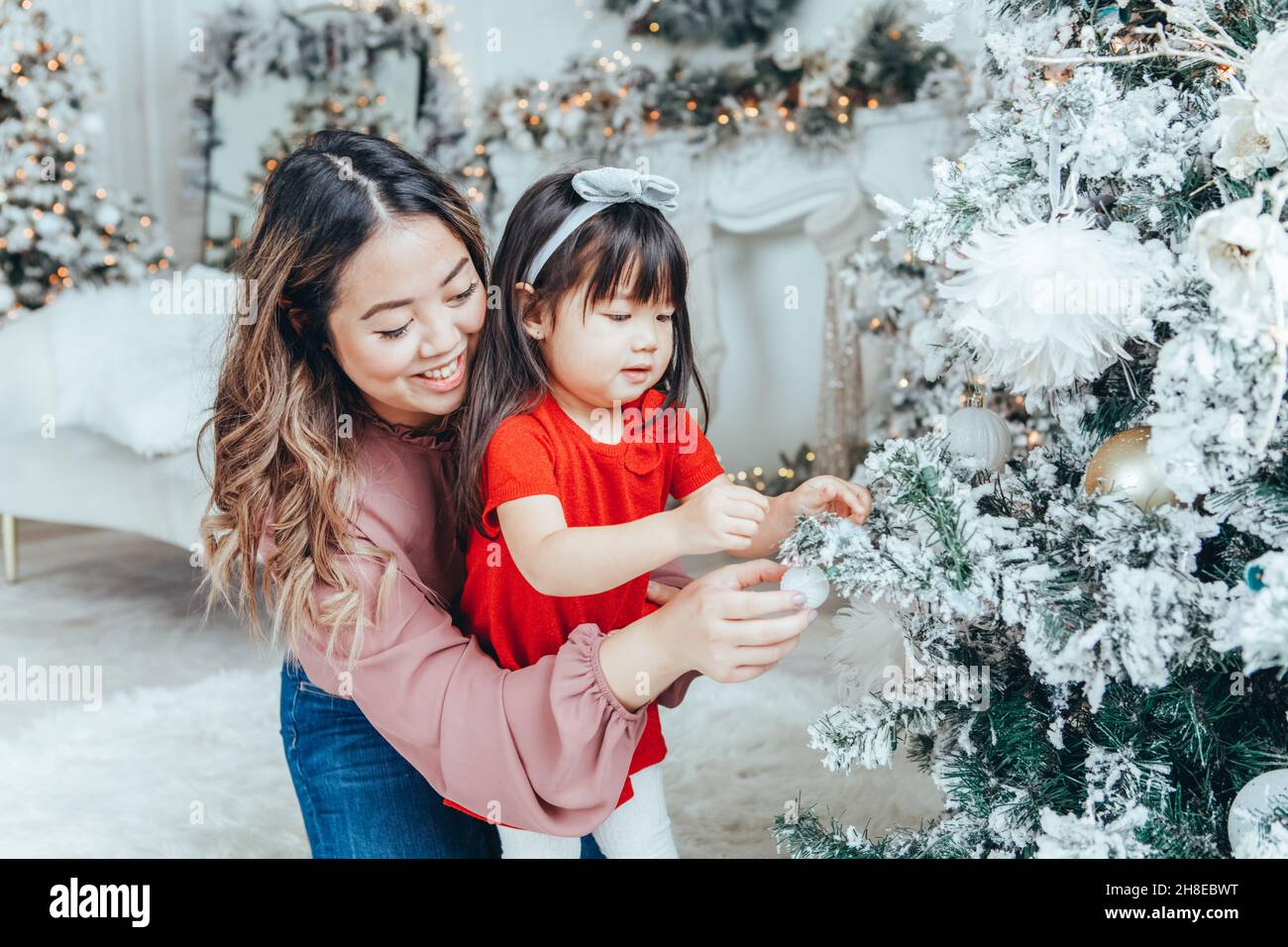 Asian family mother with daughter toddler girl celebrating Christmas or New Year. Mixed race mom, daughter decorating Christmas tree at home. Winter h Stock Photo
