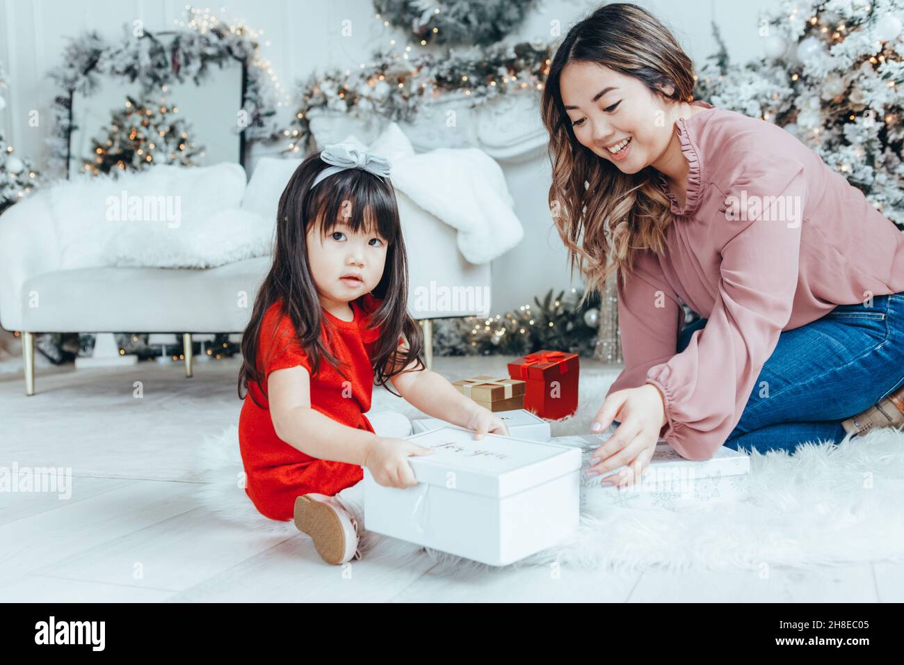 Asian family mother giving gift box with present to daughter toddler girl celebrating Christmas or New Year. Mixed race mom sitting on floor with daug Stock Photo