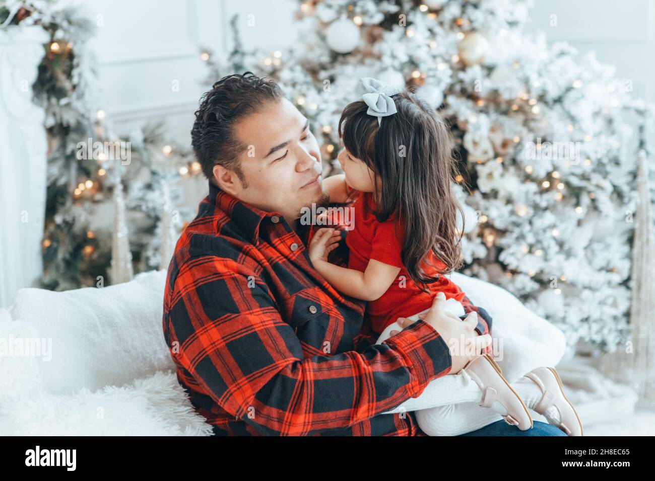 Asian family father dad hugging with daughter toddler girl celebrating Christmas or New Year. Mixed race dad man with kid sitting on couch sofa and cu Stock Photo
