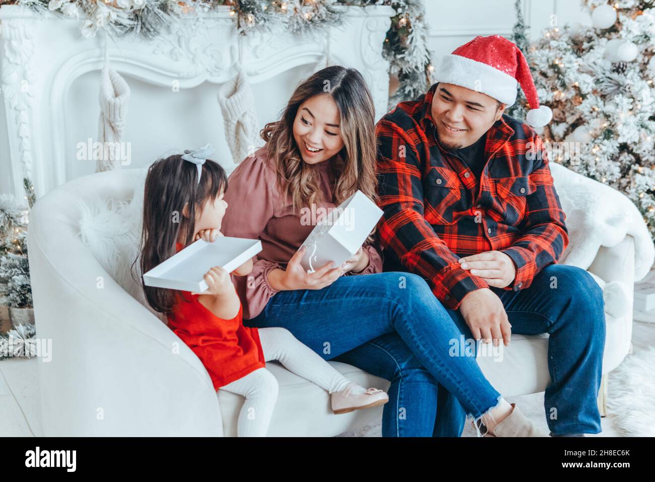 Asian family father, mother giving gift box with present to daughter toddler girl celebrating Christmas or New Year. Mixed race mom, dad, daughter sit Stock Photo