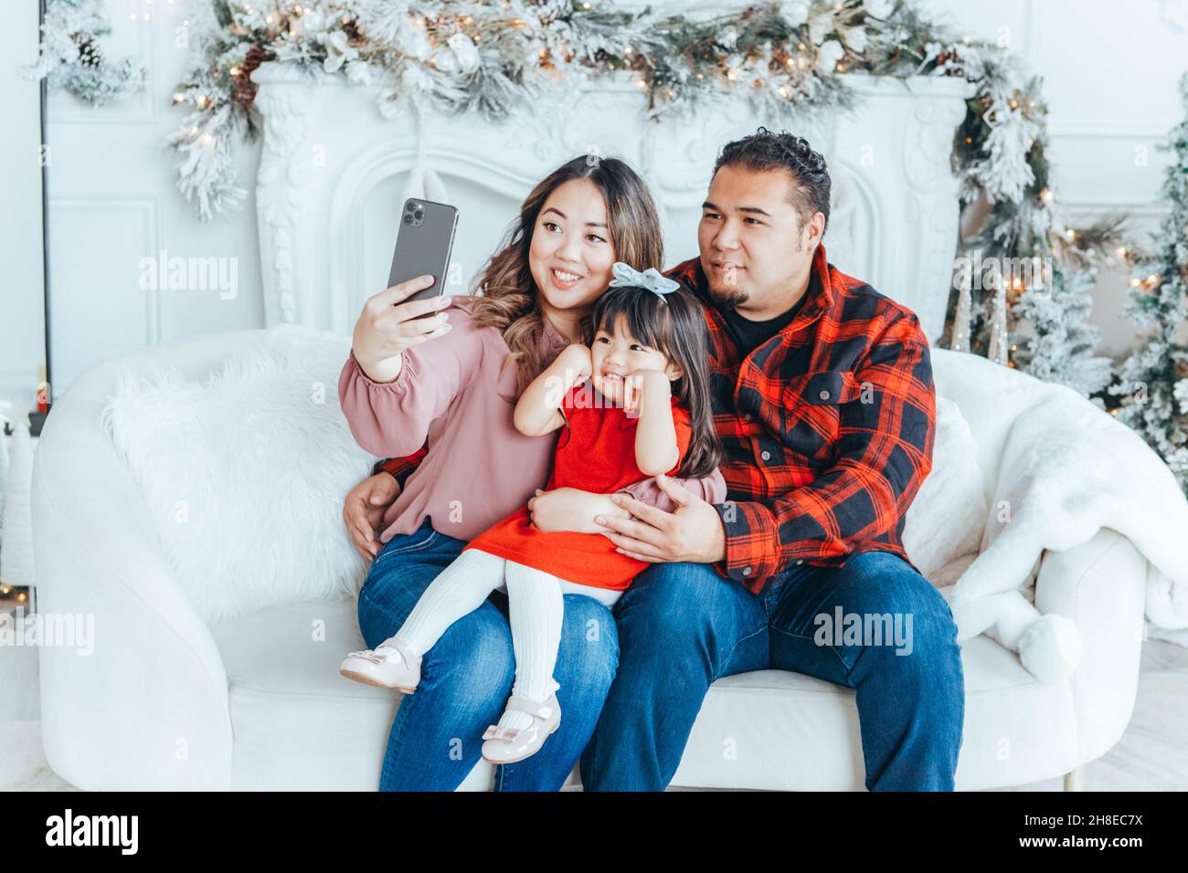Asian family father, mother with daughter toddler girl celebrating Christmas or New Year. Mom, dad, daughter sitting on couch at home and taking selfi Stock Photo