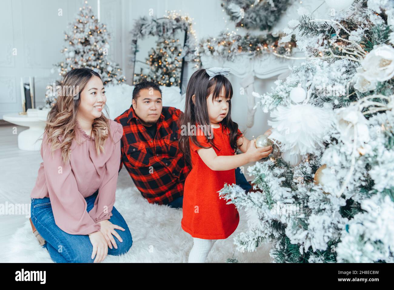 Asian family father, mother with daughter toddler girl celebrating Christmas or New Year. Mixed race mom, dad, daughter decorating Christas tree at ho Stock Photo