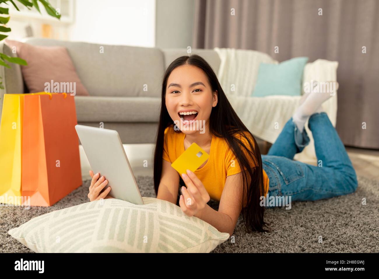 Online sales and discounts. Surprised asian lady lying on floor with tablet and credit card, emotionally reacting sale Stock Photo
