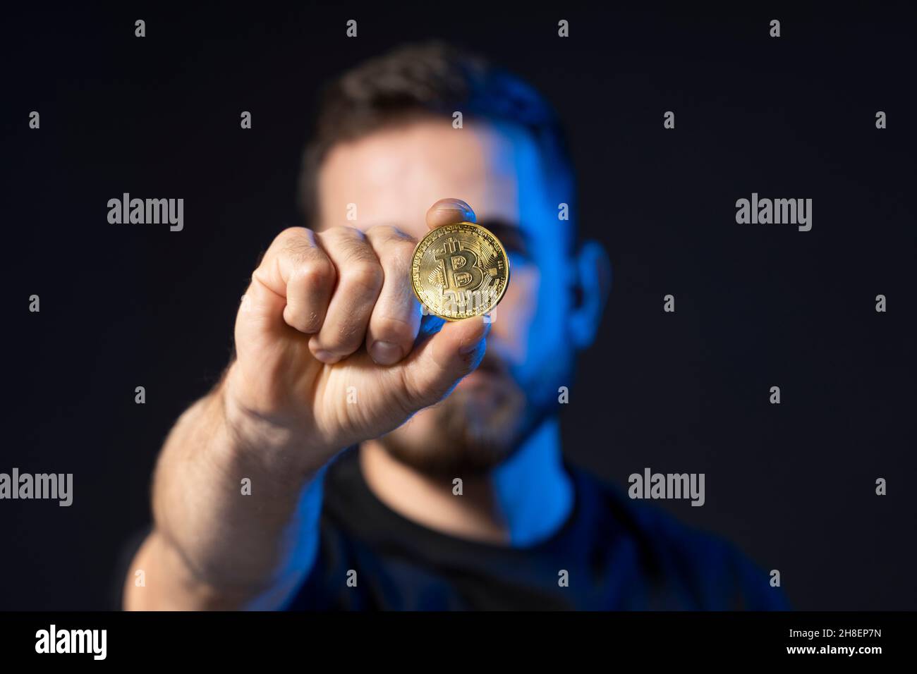Mature bearded man wearing in black t-shirt holding gold bitcoin in hand and standing isolated on black background. Stock Photo
