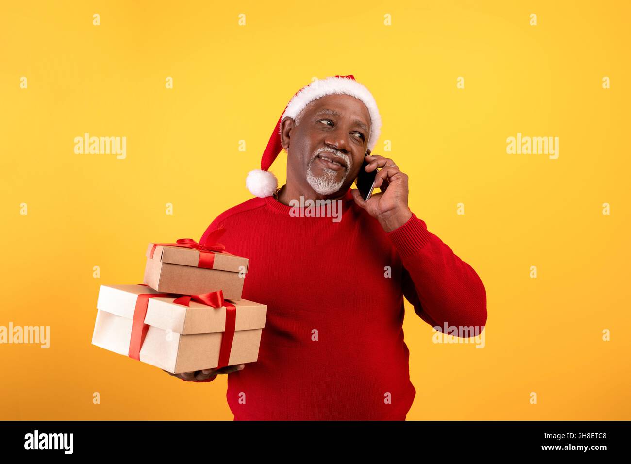 Senior black man holding Xmas gift boxes, wearing Santa hat, speaking on smartphone on orange studio background Stock Photo