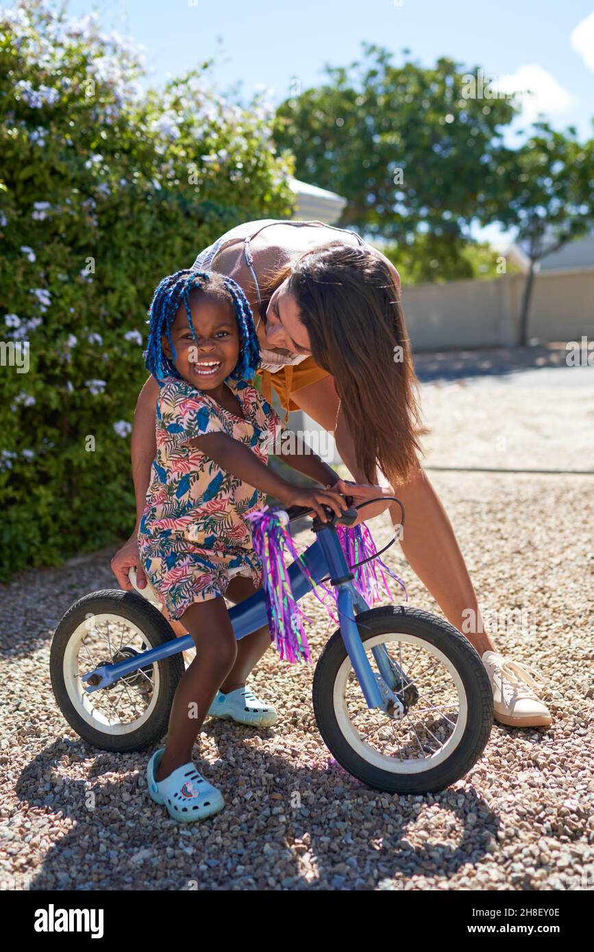 Mother kissing cute toddler daughter on bicycle in sunny driveway Stock Photo