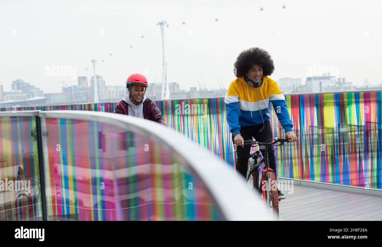 Teen friends riding bicycles on modern footbridge in city, London, UK Stock Photo