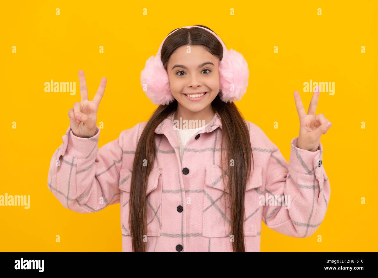 Happy girl child smile in warm winter fur earmuffs gesturing double V signs hand gesture, victory Stock Photo