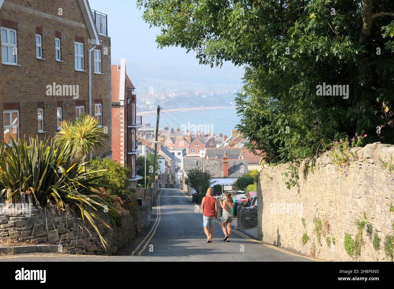 Swanage, Dorset, UK.  A couple walking down a steep hill on a summer's morning towards the town centre with views over the rooftops to Swanage Bay. Stock Photo
