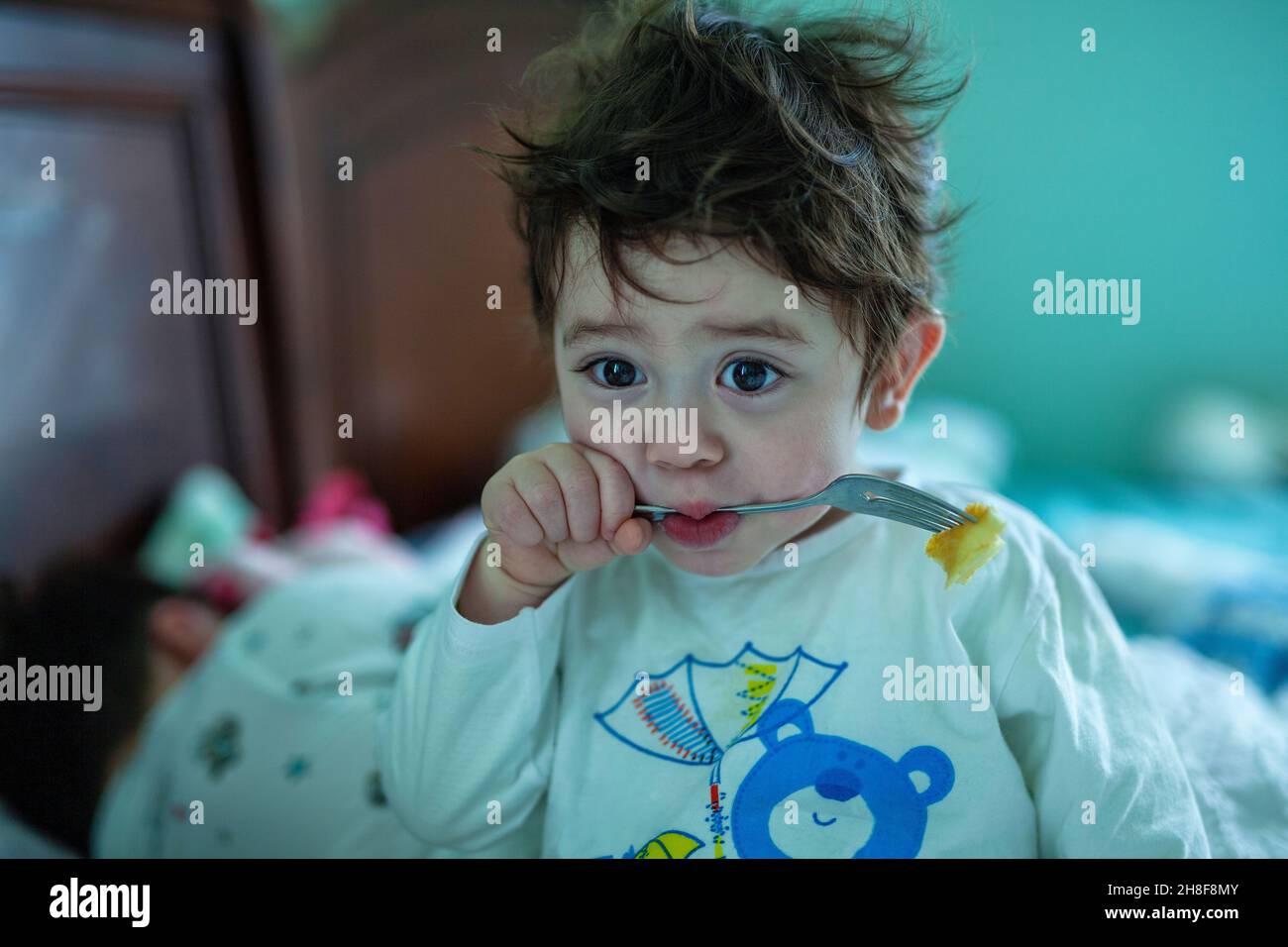 Little Boy with large brown eyes and messy brown hair wearing white pajamas with a blue bear eating a pancake with a fork Stock Photo