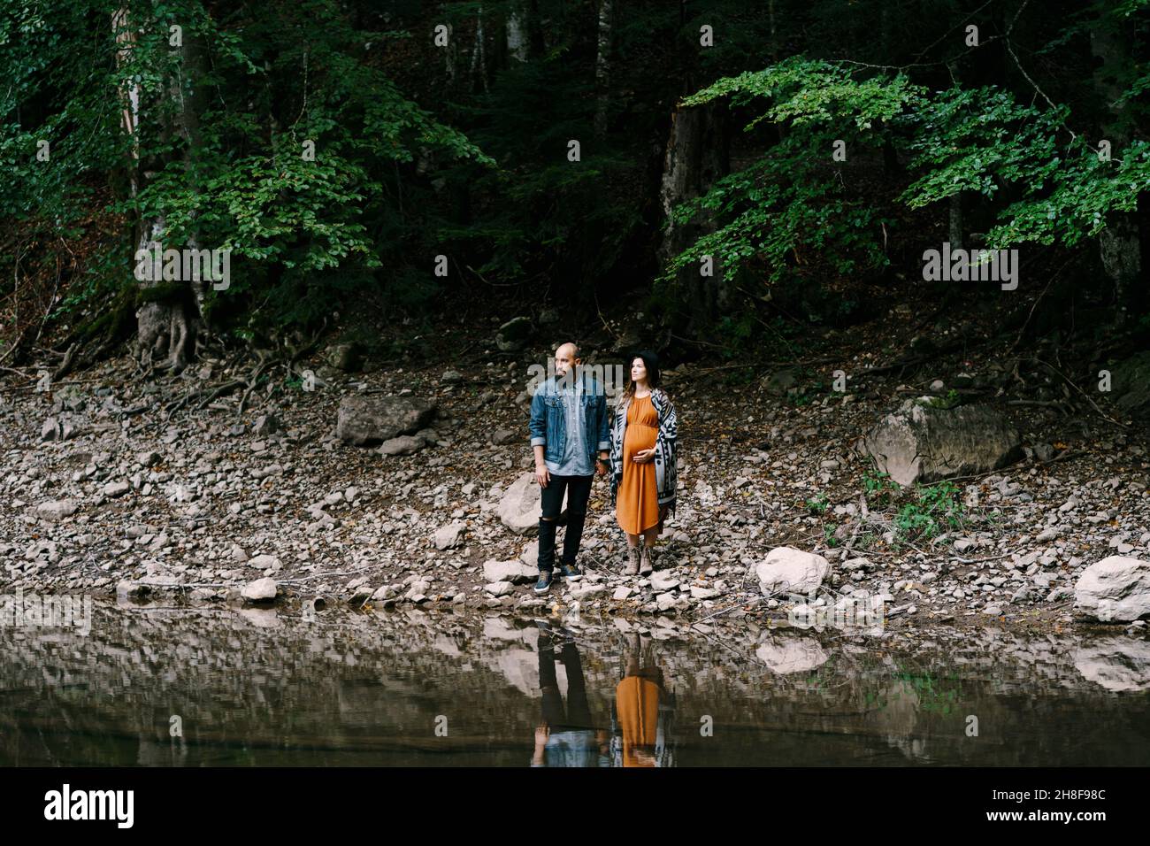 Pregnant woman with man stands on a rocky slope by a lake surrounded by ...