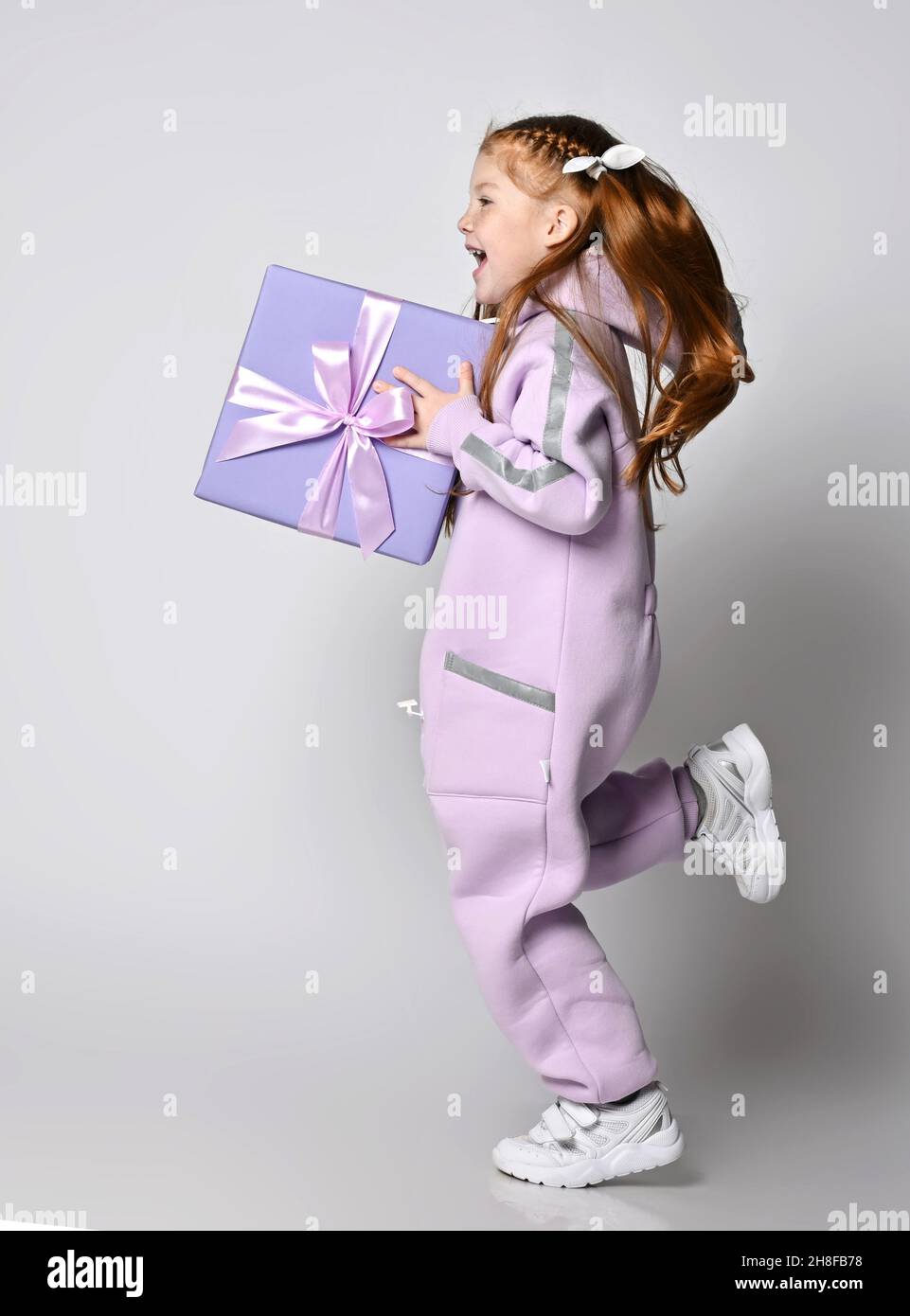 Frolic laughing kid girl in pink jumpsuit is running holding big blue gift box, birthday present in hands. Side view Stock Photo