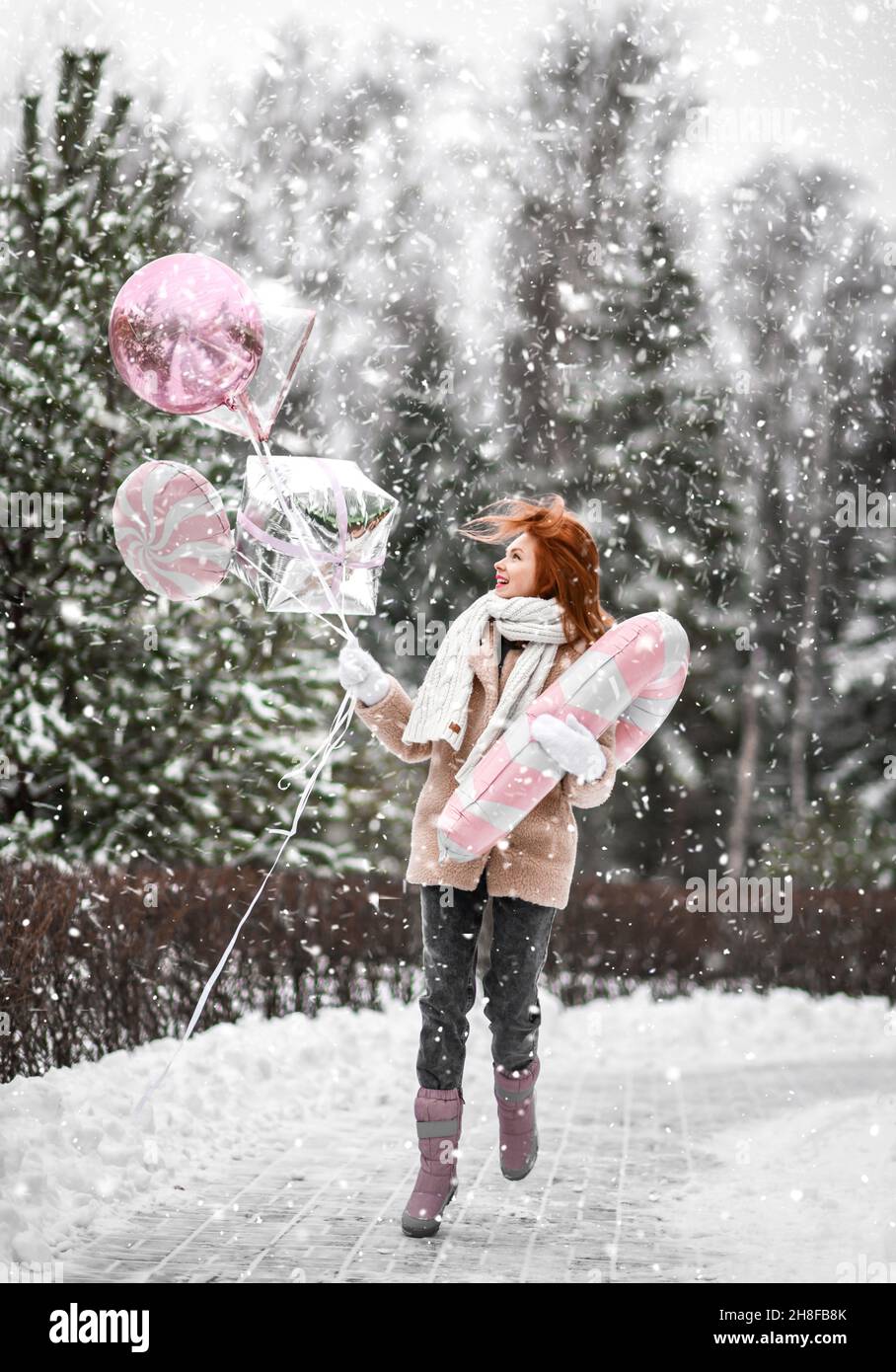 Young happt pretty woman in stylish fur coat and mittens walking with toy magic wand and glitter balloons in hands in winter park Stock Photo