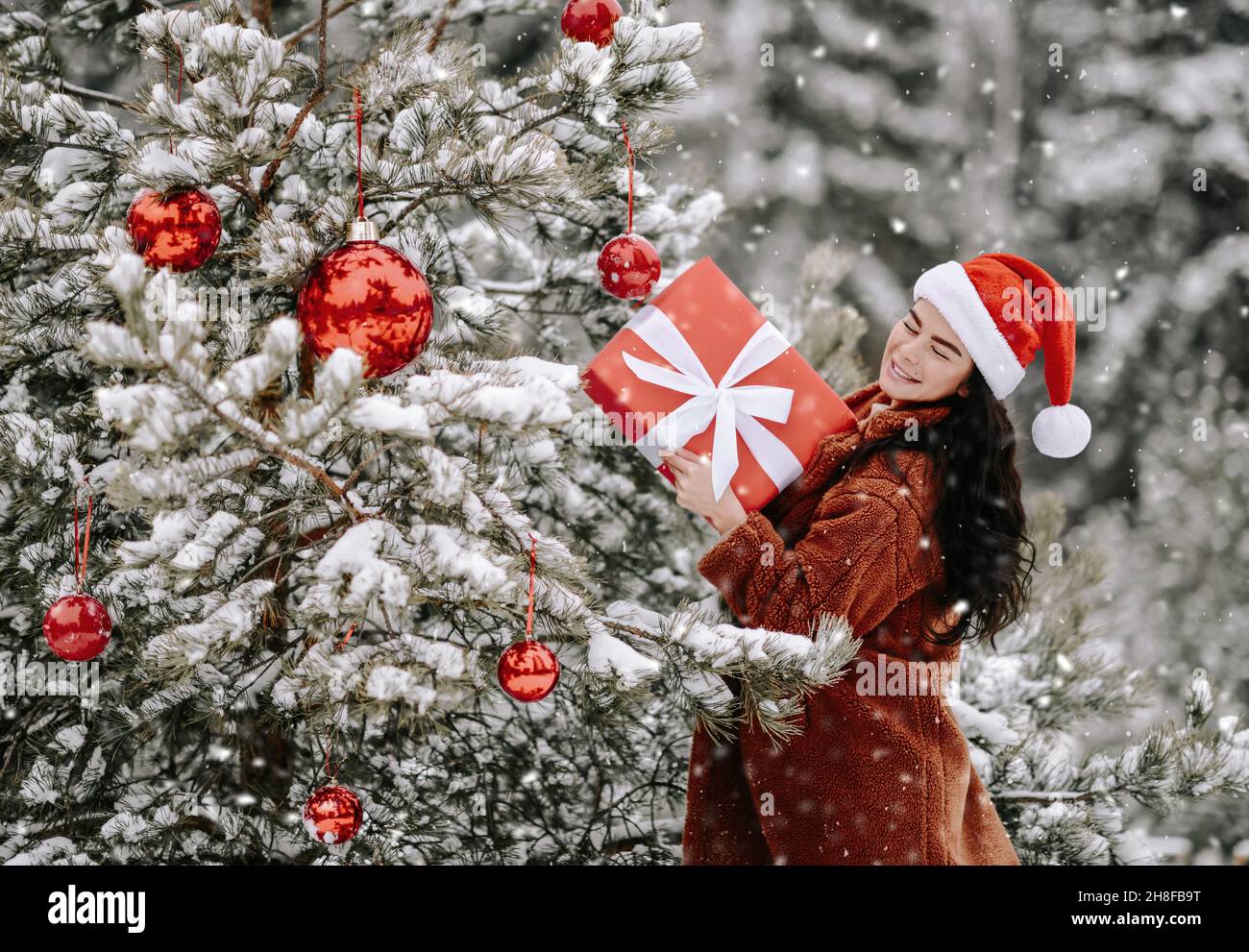 Young smiling beautiful woman in stylish fur coat and santa claus hat hold holiday gift box in hands Stock Photo