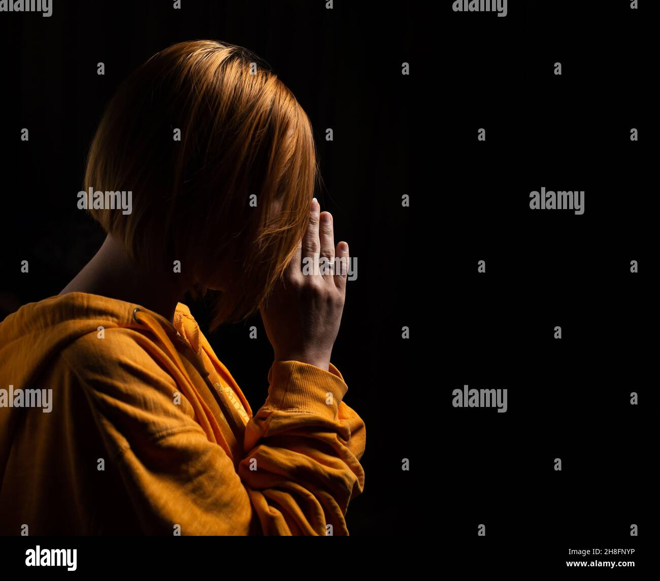 Woman praying with her head bowed with folded palms on a black background. Stock Photo