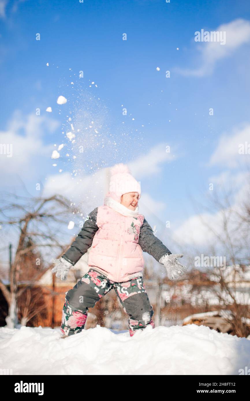 Girl is playing snowballs against blue sky. Child in warm winter suit has fun, plays and jumps on snowdrifts, rides snow slide while walking in nature Stock Photo