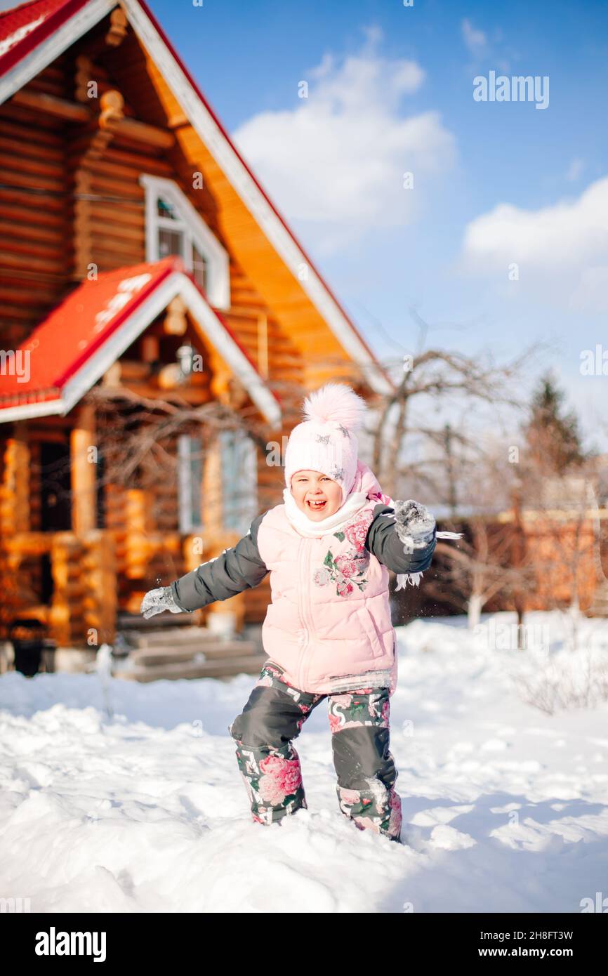 Little girl is playing on background of cottage. Child having fun on sunny winter day in backyard near wooden house, playing snowballs, jumping in Stock Photo