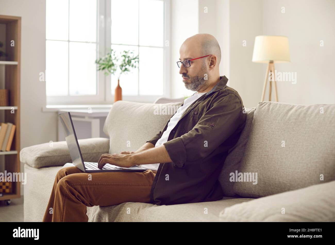 Serious adult man sitting on couch at home and working on modern laptop computer Stock Photo