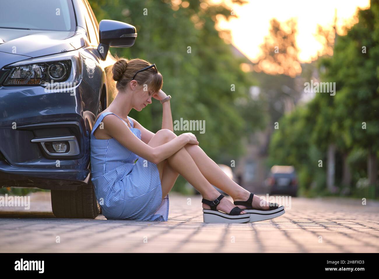 Young woman driver sitting beside her broken car waiting for help. Vehicle problems concept. Stock Photo