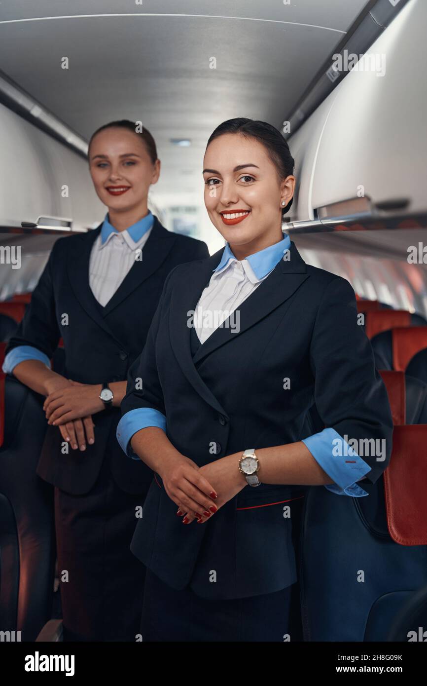 Two stewardesses standing on airplane aisle with folded hands Stock Photo