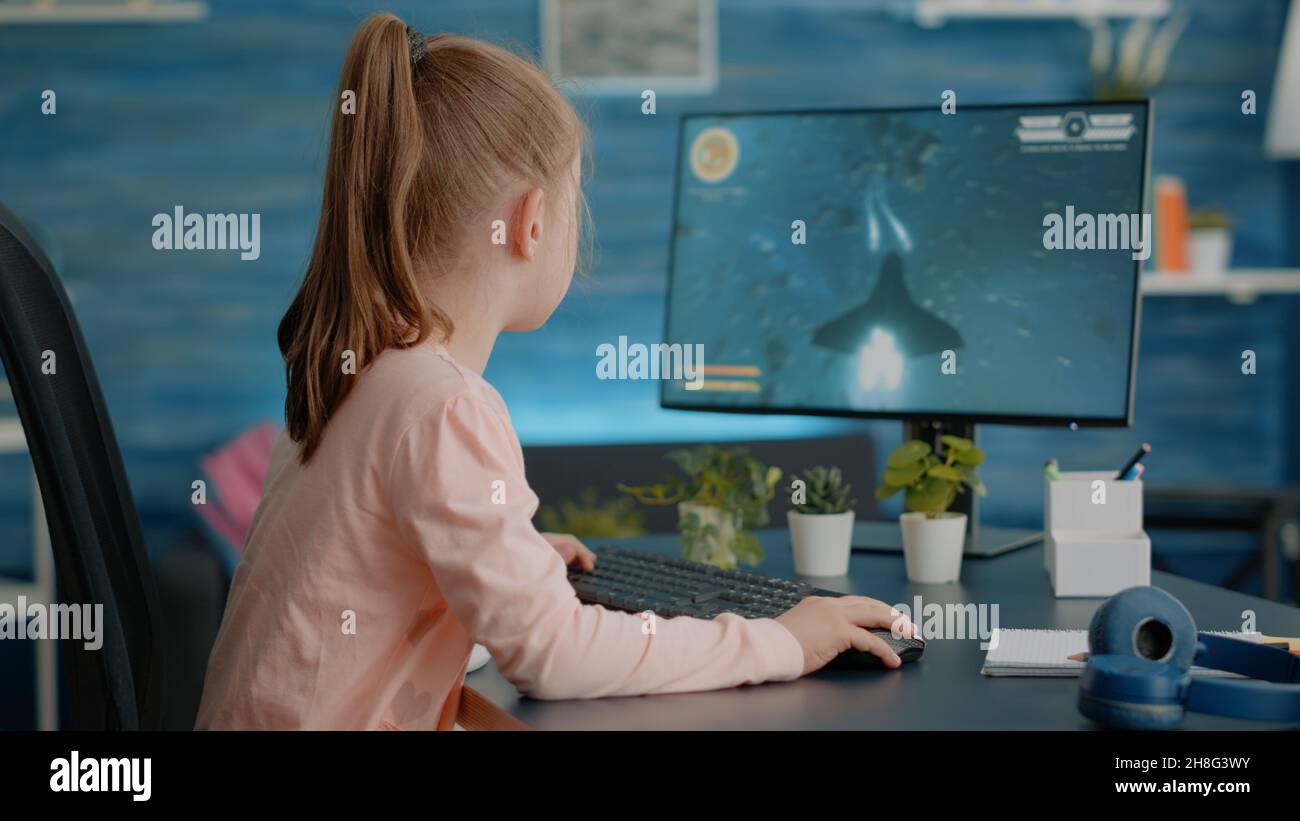 Schoolgirl using computer to play video games for entertainment at home. Child playing action shooting game with monitor and keyboard, enjoying fun activity after doing homework. Stock Photo