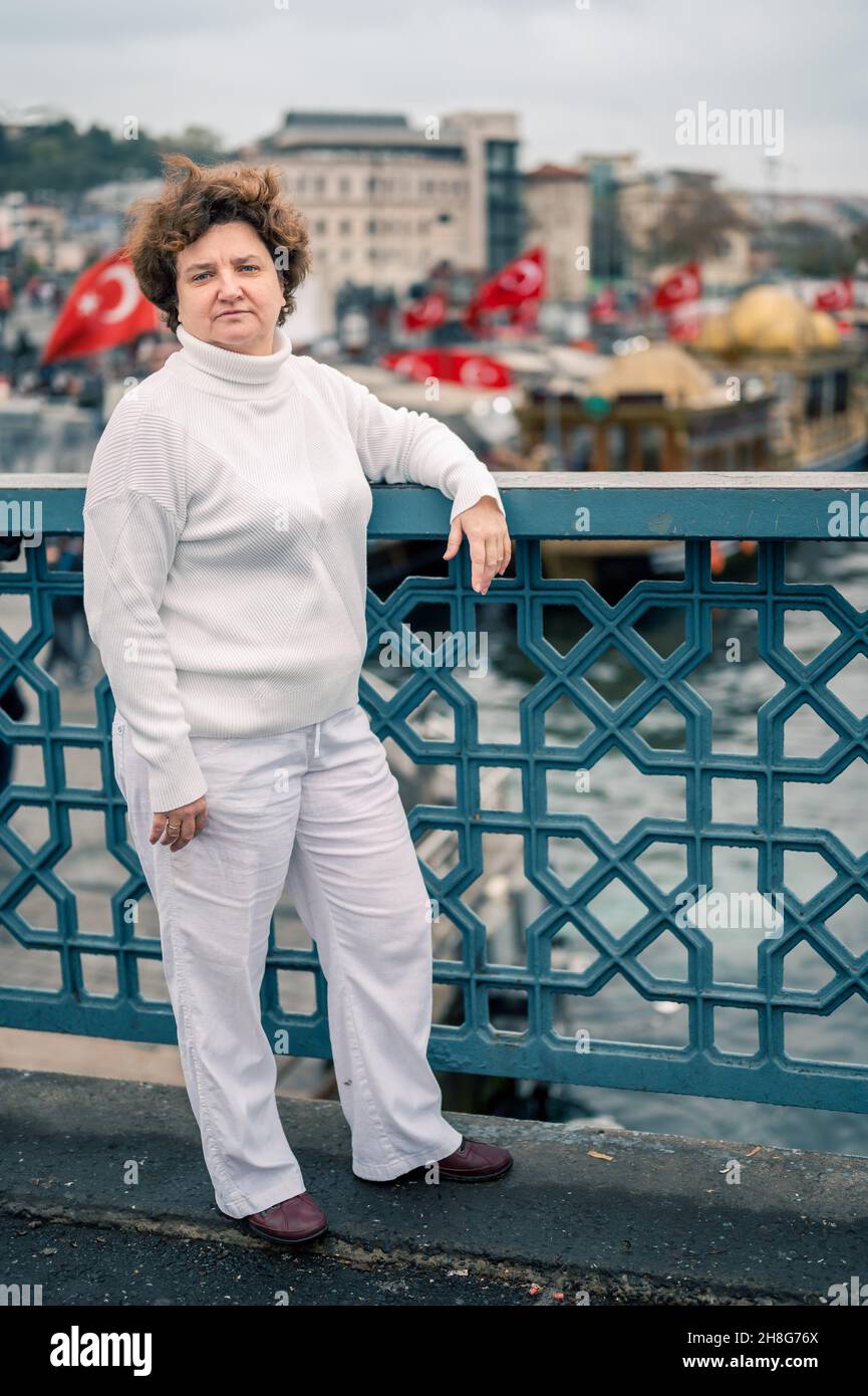 Ederly woman in white clothes on Galata bridge on cloudy day. turkish flags are flying in background. a 50-year-old woman, short, looks into camera , Stock Photo