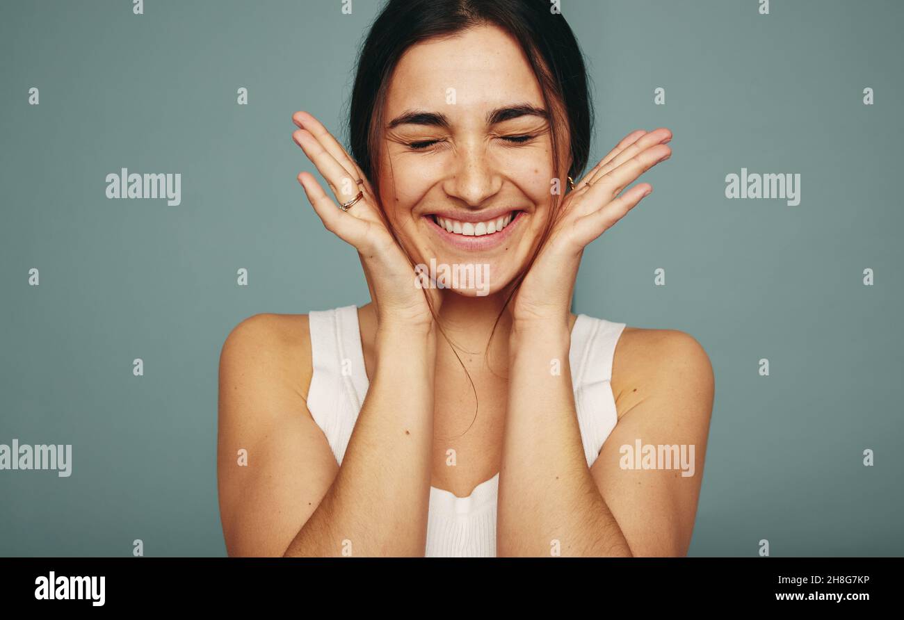 Excited young woman smiling with her eyes closed in a studio. Happy young woman embracing her face while standing alone against a studio background. Stock Photo
