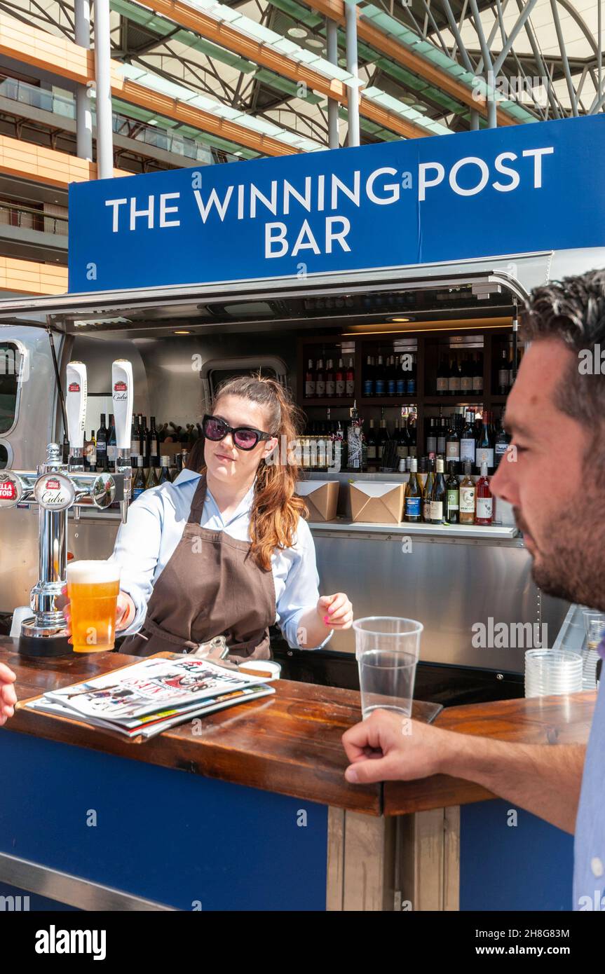 The Winning Post Bar at Royal Ascot racecourse, Berkshire, UK. Barmaid serving a customer with a draught pint of lager. Serving alcohol on sunny day Stock Photo