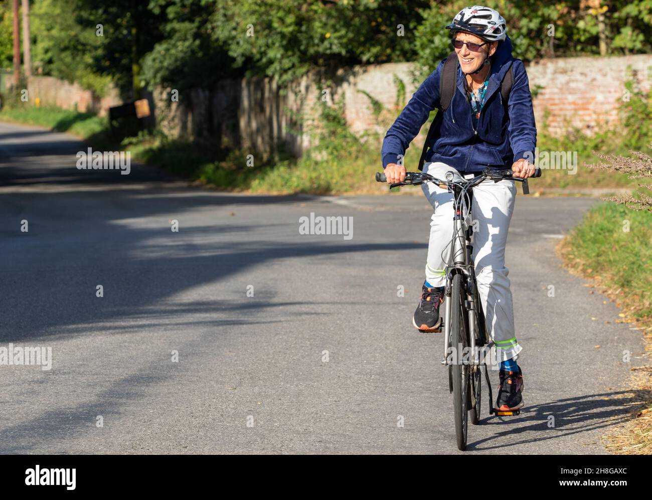 On a sunny day a woman rides her bicycle slightly uphill on a quiet Suffolk country road Stock Photo