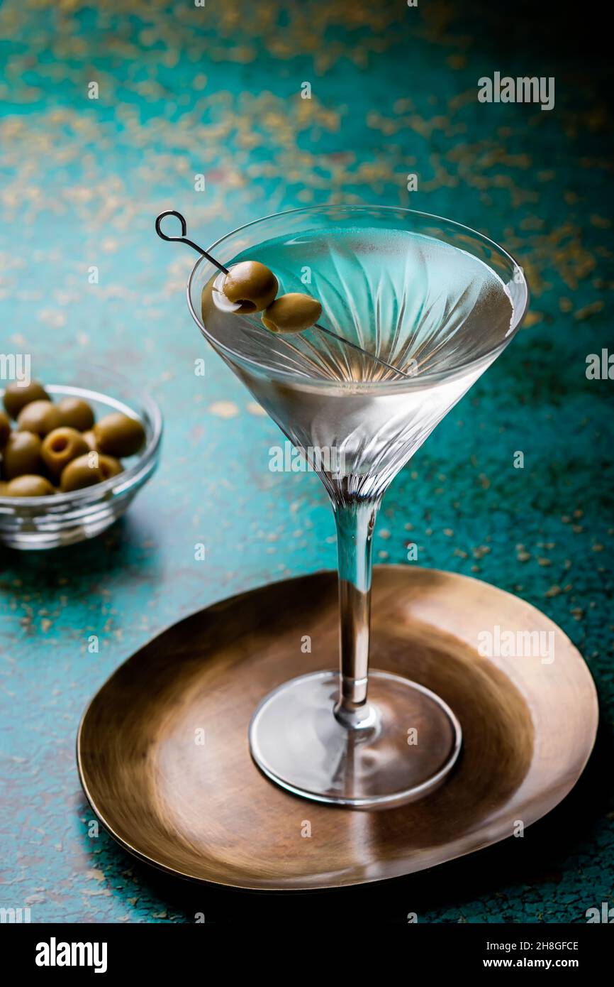 Dirty martini cocktail closeup in back light with olives garnish on green textured table closeup with plate with olives in background Stock Photo