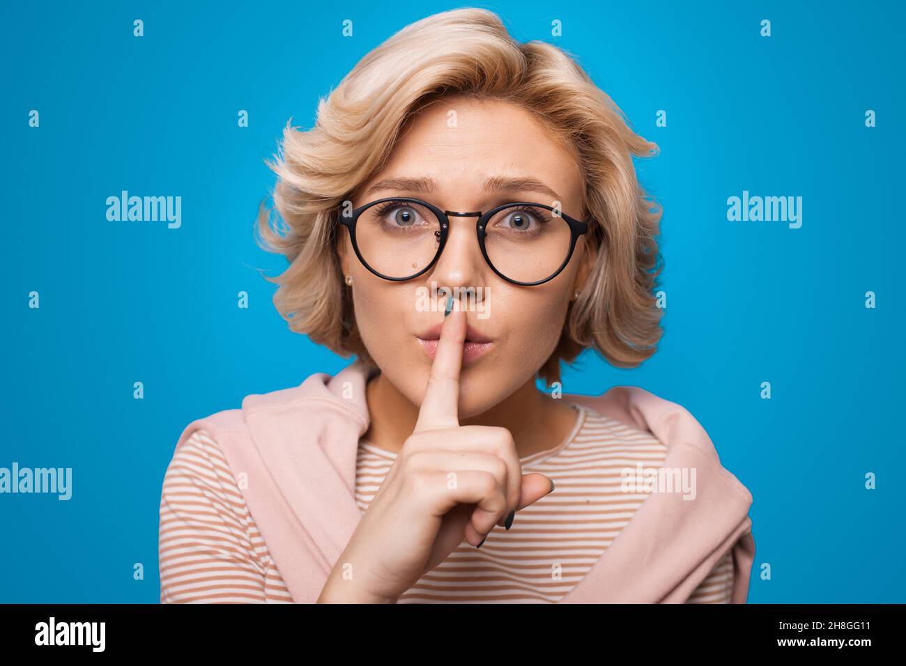 Surprised blonde haired woman makes silence gesture presses index finger over lips isolated over blue background. Shhh sign. Stock Photo
