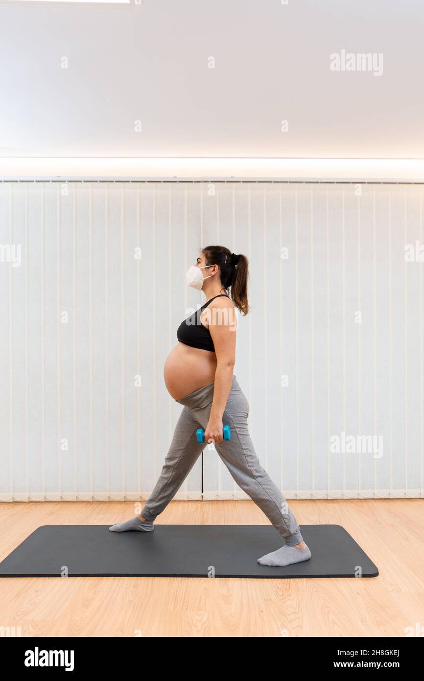 A pregnant woman with face mask standing doing dumbbell exercises at home Stock Photo