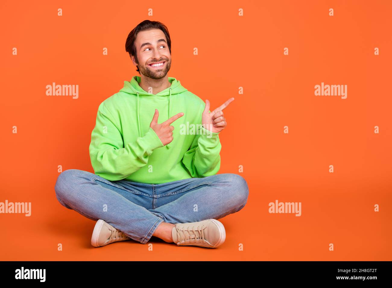 Full size photo of brunet young guy sit index promo wear green hoodie jeans shoes isolated on orange color background Stock Photo