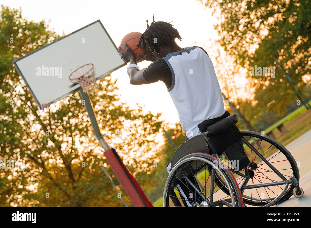 Young athlete on wheelchair throwing ball in basketball basket, sunset