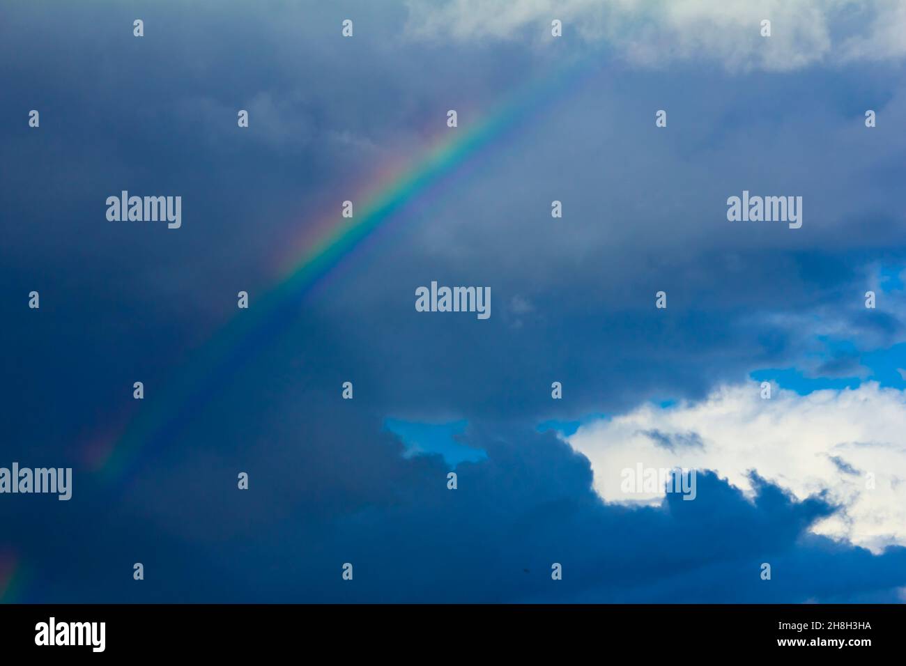 Colorful rainbow with dark storm clouds against a background of blue summer sky. Concept of change, hope. Stock Photo