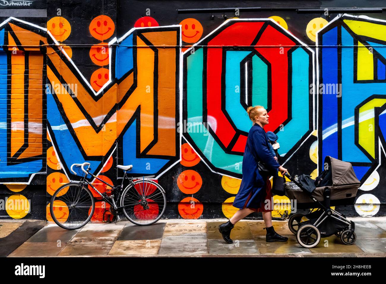 A Young Woman Walks Past Some Street Art/Graffiti Pushing A Pushchair, Shoreditch, London, UK. Stock Photo