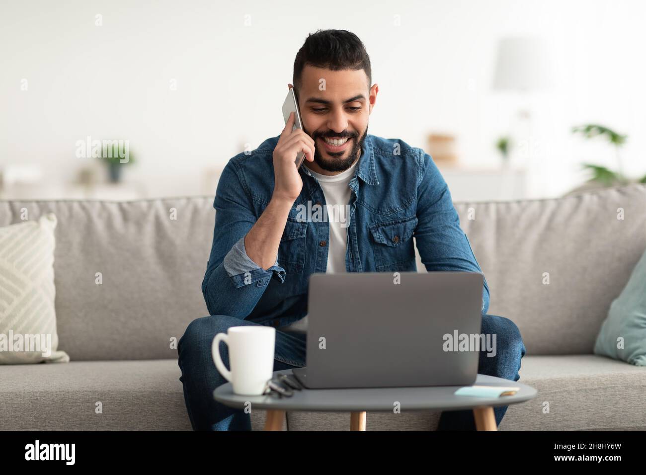 Work from home. Positive young Arab guy talking on mobile phone while using laptop computer indoors Stock Photo