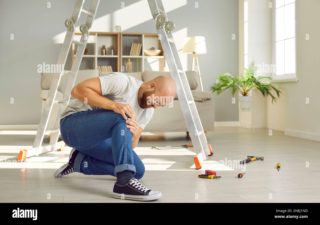 Young man who just fell off ladder is sitting on floor and hugging his hurt knee Stock Photo